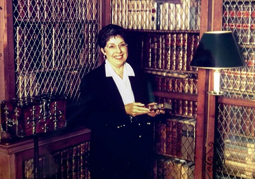 Jane Millgate stands smiling in front of shelves of books at the Walter Scott library