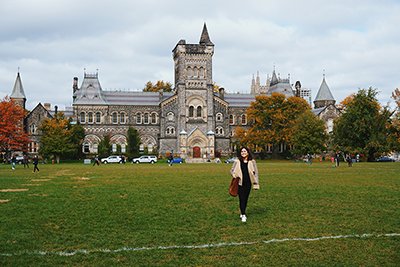 A smiling student walks across U of T's front campus lawn. University College is behind her.