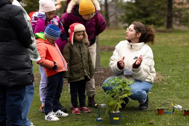 Children looking at a person speaking beside a small plant in a pot.