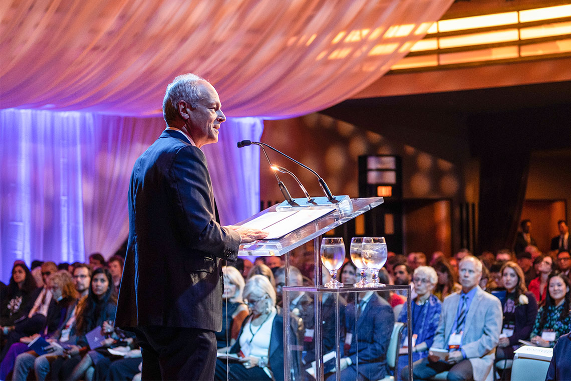 President Meric Gertler stands at a podium welcoming a crowd of over 100 people.