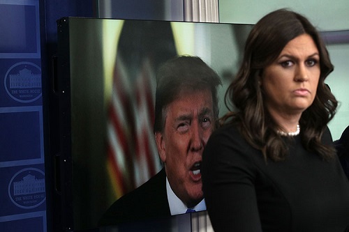  Sarah Sanders listens during a daily news briefing at the White House with an image of Donald Trump on a computer screen.