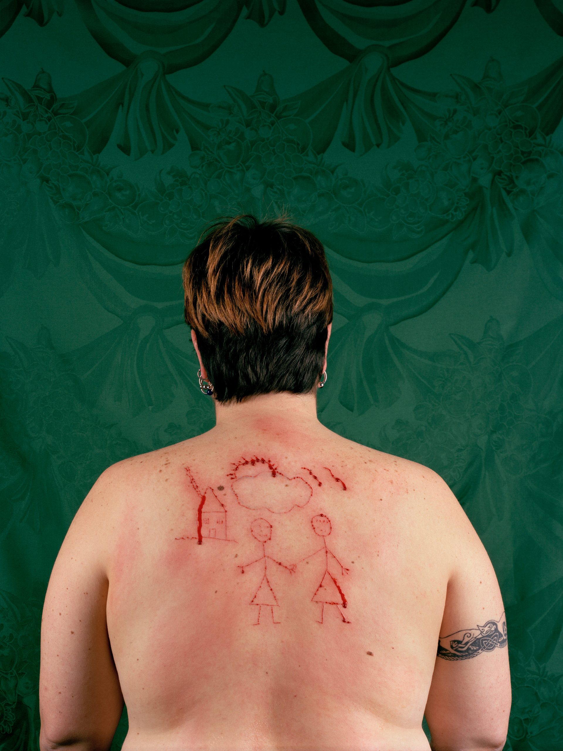 A woman's back with a stick figure drawing of two women and a house etched into her skin