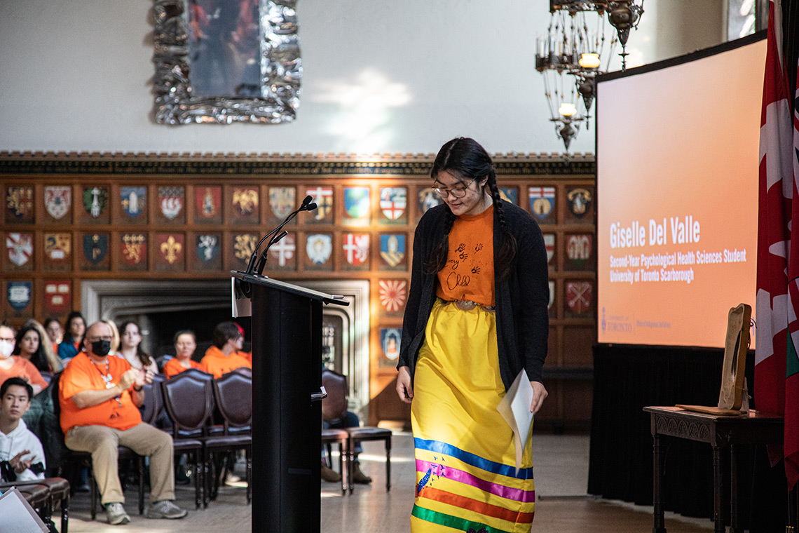 Giselle Del Valle at a podium in a yellow skirt and orange shirt