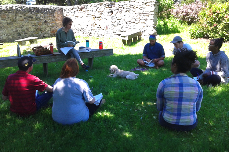 Students sit in a circle on grass and listen to Professor Andrea Most's lecture. A dog is lying in the middle of them.