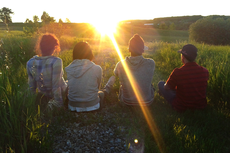 Four students sit on the grass, facing the sunset on the horizon