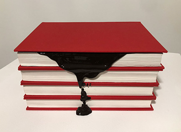 A stack of red books, the top book has black paint dripping from the pages.