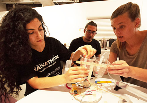 Three students connect two cups and sticks during an experiment.