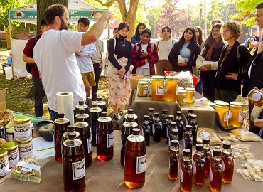 In foreground a table covered in jars of honey, behind table a group of students listening to a man speaking