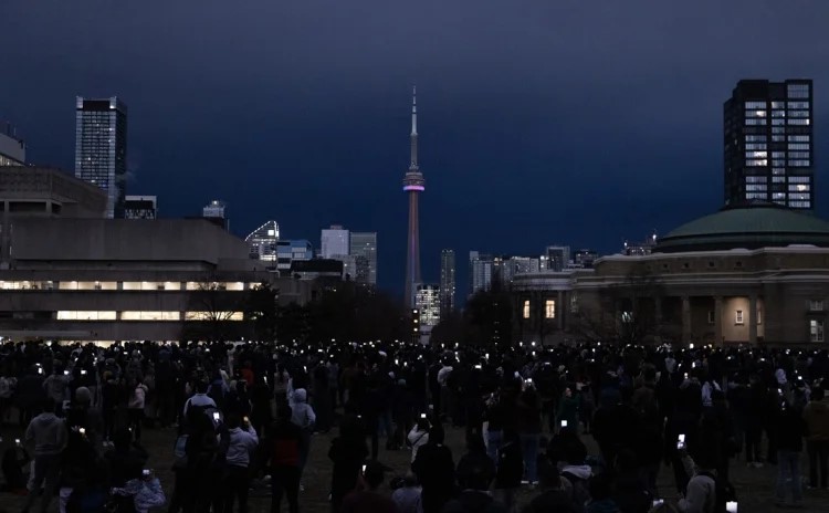 A view of the CN Tower under a dark sky and hundreds of people standing in the middle of U of T's St. George campus, looking at the total eclipse through their phones.
