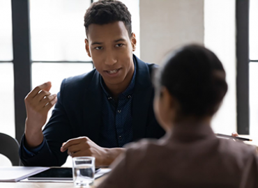 A person speaking with another person at a desk.