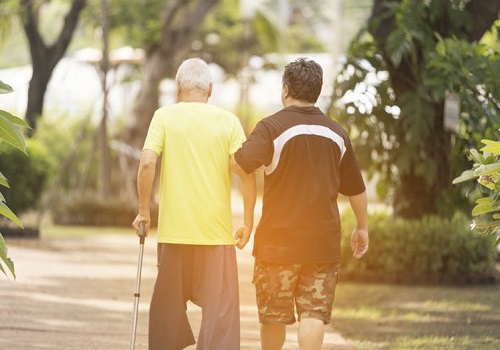 An elderly man walks with a cane. A middle-aged man walks with him, holding the elderly man's arm