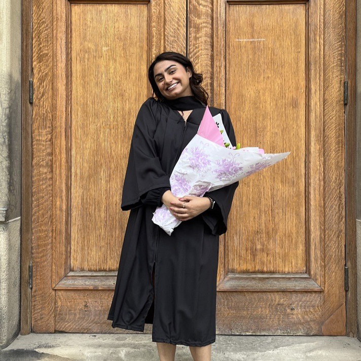 Mariam standing in front of wooden doors in a convocation gown holding a bouquet of flowers