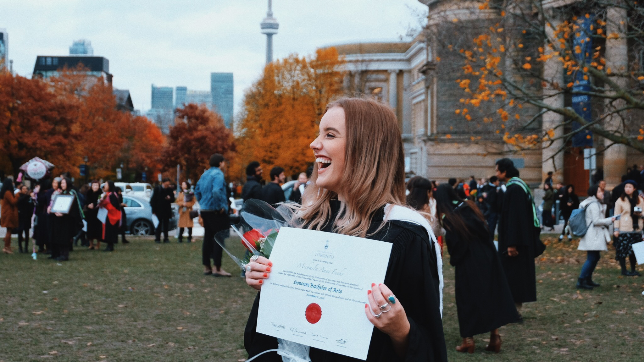 Student in graduation robe standing in front of Convocation Hall holding up diploma 