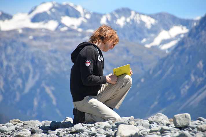 A student kneels at the top of the mountain in New Zealand with a book