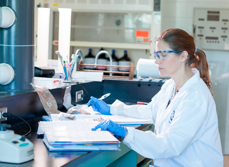 A woman, wearing a lab coat and safety glasses, writes in her notebook inside a lab.