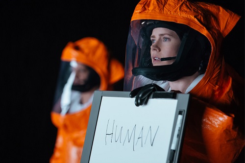 Louise Banks, played by Amy Adams, in an orange spacesuit, attempting to communicate with aliens, first by introducing a white board that says human.