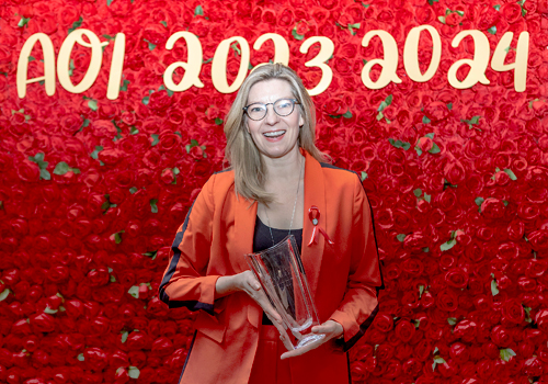 Maev Beaty holding a glass award, roses and the text "AOI 2023 2024" in the background.