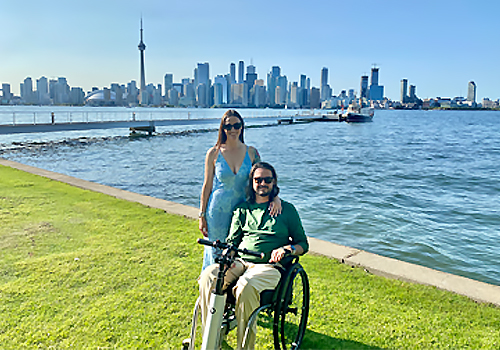 Beau outside with his partner, Meghan beside Lake Ontario with the CN tower in the background on a sunny summer day.