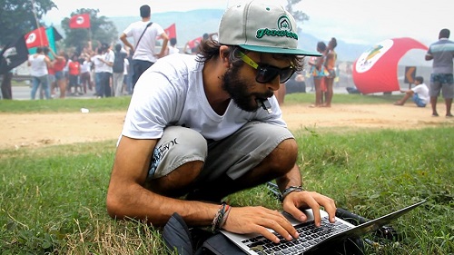 A casually dressed man, crouched down at an outdoor event, typing on a laptop, while holding a flash drive in his mouth.