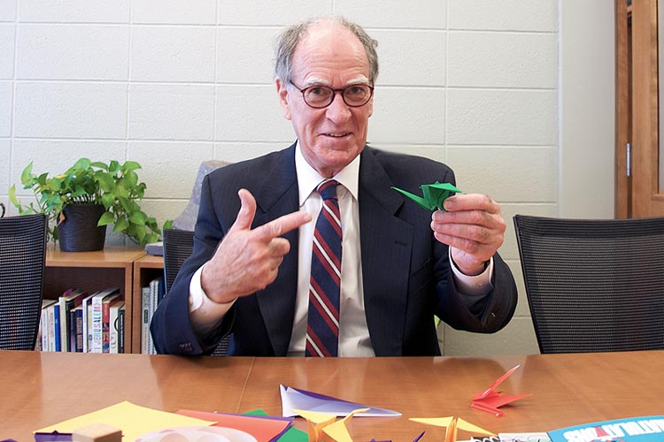 Arts & Science Dean David Cameron, sitting at a table, pointing at a green paper crane he is holding with his left hand.