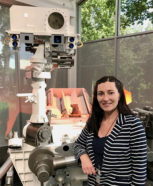 Carrie Bridge standing beside a large piece of machinery called the curiosity rover.