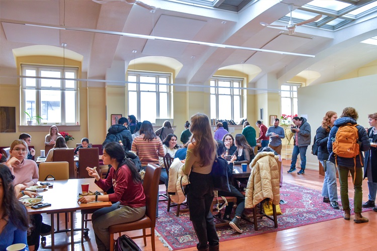 A group of student and faculty in a room with big windows standing in groups and eating at tables