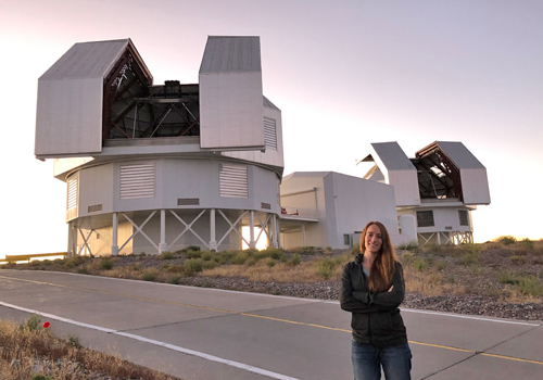  Maria Drout standing in front of a large telescope at sunset.