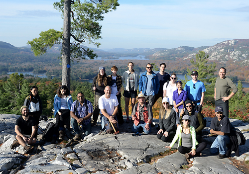 A group of students standing on a cliff with a hilled forest landscape and lakes in the background.