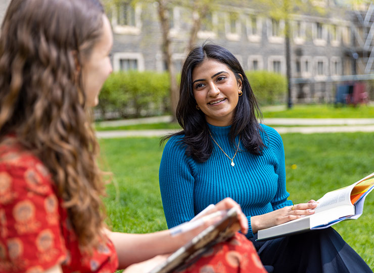 Two students sit together in a grassy quad with textbooks in hand