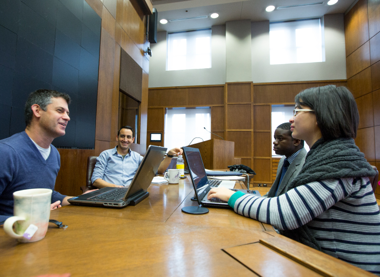 A professor has a discussion with a group of graduate students inside a meeting room.