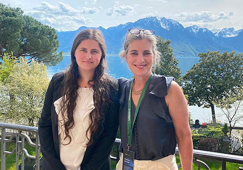 Ariel Greiner and Dean Melanie standing outside on a sunny day with a beautiful scene of a mountain and lake in the background.
