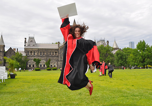 Christine Hone-Buske jumping in front of University College wearing a graduation gown and holding her diploma.