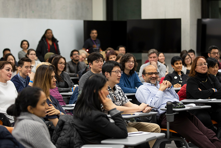 A crowd of attendees sits in a classroom. A man in the front row laughs.