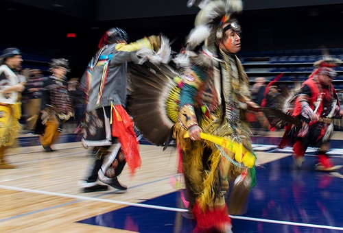 A group of First Nations people dance in colourful clothing during a powwow inside a U of T gym.