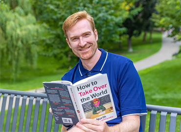 Ryan North sitting on an outdoor bench holding his book