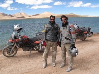 Ryan Pyle with brother Colin standing in front of their motorcycles, a large body of water, and sand dunes.