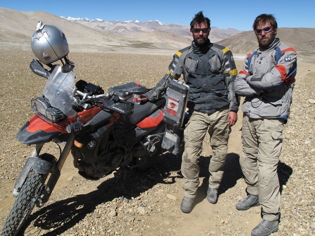 Ryan Pyle with brother Colin standing by a motorcycle in a barren landscape with snowcapped mountain in the distance.
