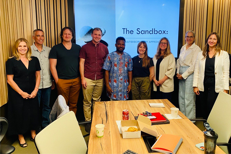 A large group of people gathered around a table with a presentation in the background that says, The Sandbox.