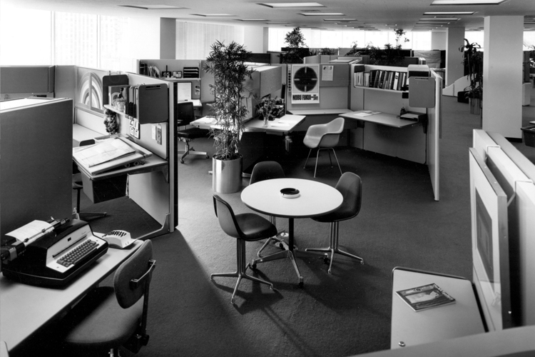 A 1960s office setting, including cubicles in a half-hexagon shape, Herman Miller chairs, a typewriter, etc.