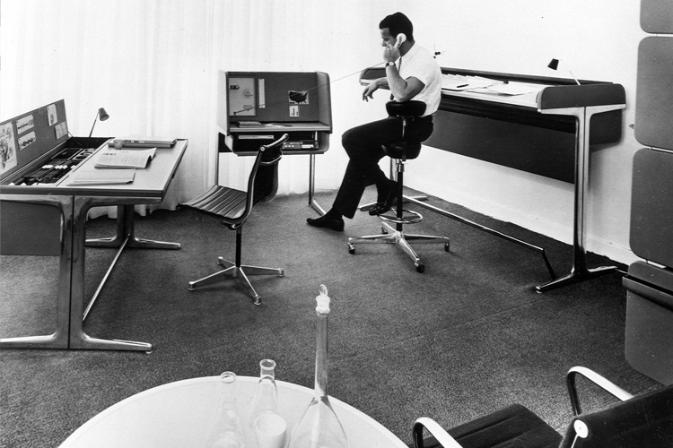 A man sits in his office in the 1960s. He is on the phone at a bar-height office chair with a tall desk behind him.
