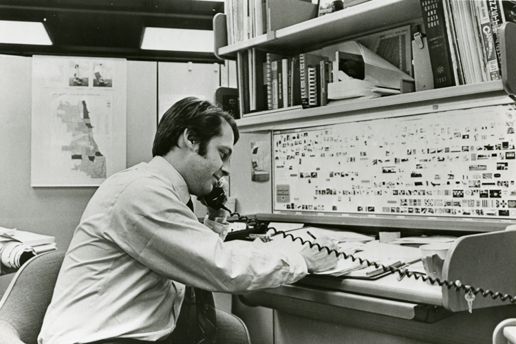 A seated man uses a phone at a small desk. There is a bookshelf above him and a map behind him.