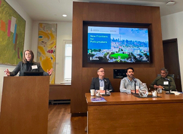 Shiri Breznitz standing at a podium and Derek Newton, Aman Thind and Deepak Ramachandran sit at table at the front of a room.