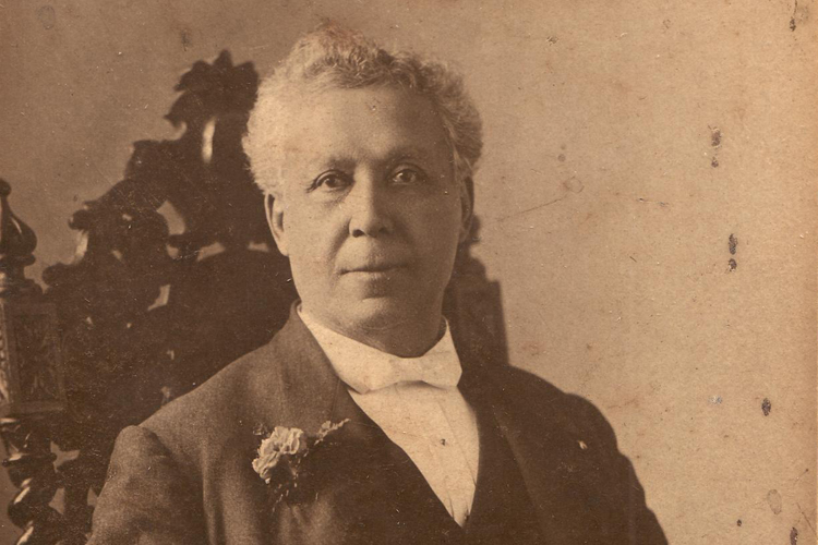 Portrait of Oronhyatekha, seated in a chair, with a flower in his buttonhole, a watch on a chain and a rolled-up publication in his left hand.