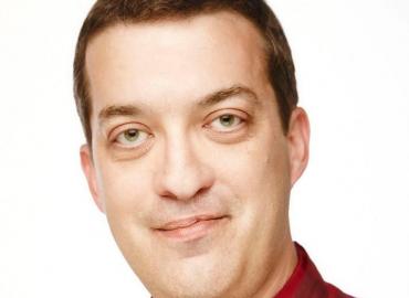 The image is a professional headshot of a man with short brown hair, light green eyes, and fair skin. He has a subtle smile and is wearing a red collared shirt. The background is plain white, providing a clean and well-lit portrait. The lighting is even, 