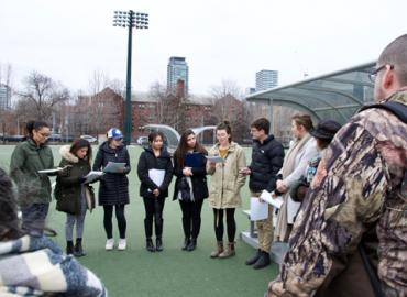 Students stand in a circle on U of T Back Campus&amp;#039;s turf field, telling stories.
