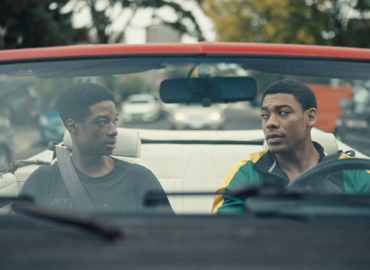 Two men seated in a convertible looking at one another.