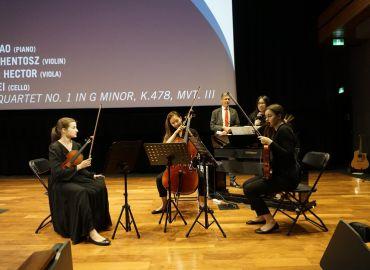 Three students seated on stage, two are playing violins and one is playing the cello.