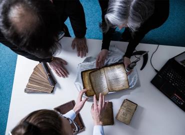 A group of three people around a table observing an old book.