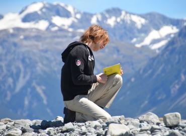 Student at the top of a mountain kneeling down with a book in their lap