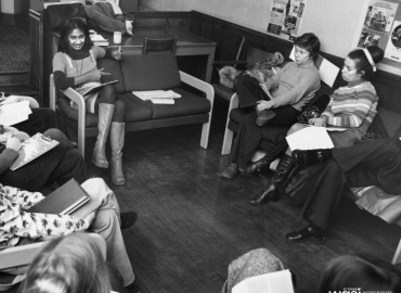 Black and white photograph of a group of women sitting in chairs in a circle. They have notebooks and are looking at a woman who is talking.